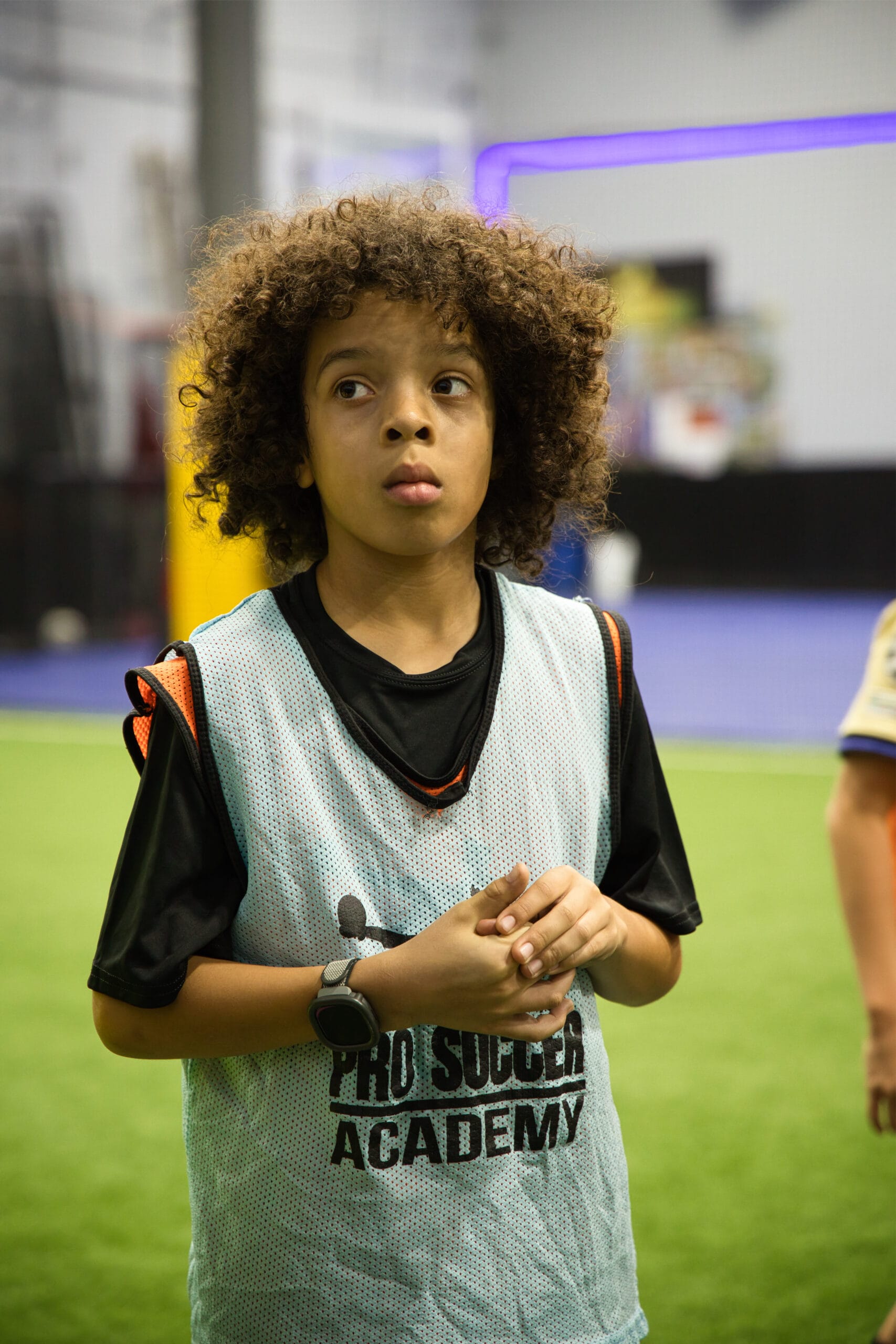 4Q8A0245 Youth soccer player with curly hair in blue Pro Soccer Academy pinnie listening attentively during indoor training session.