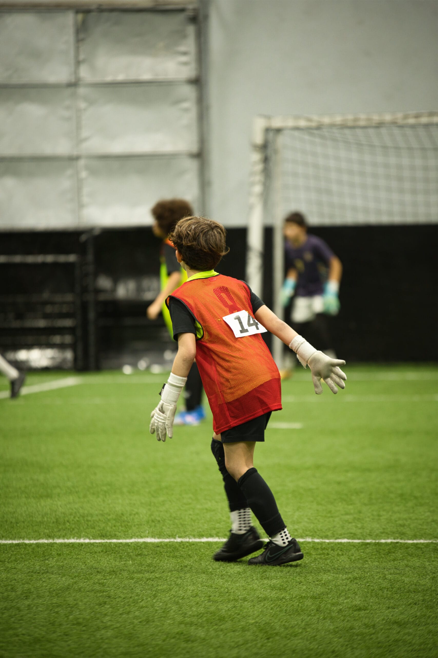 4Q8A0261 Youth goalkeeper wearing red pinnie number 14 and gloves moving into defensive position during indoor soccer match.