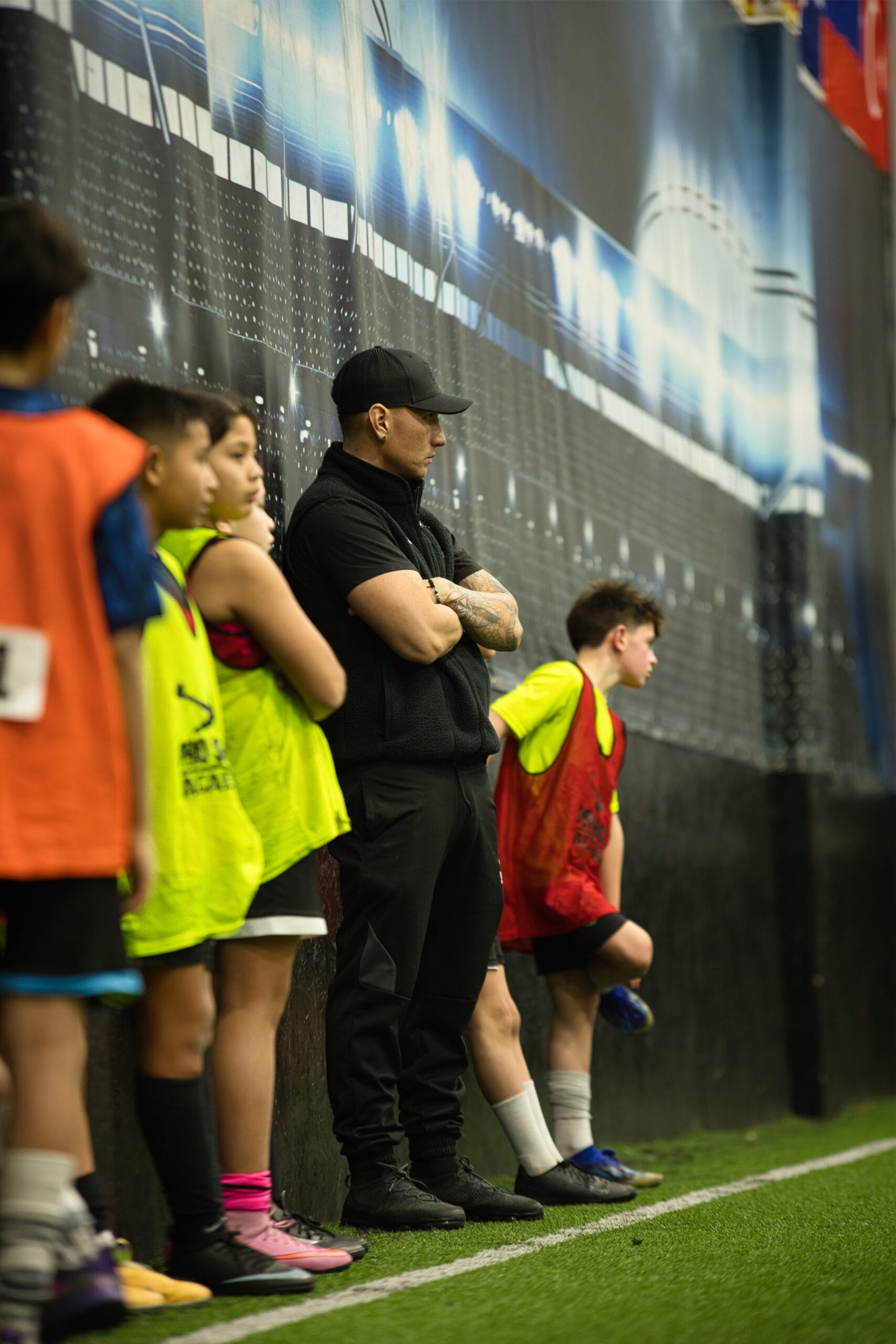 4Q8A0291 Soccer coach with arms crossed observing youth players lined up along indoor training facility wall.