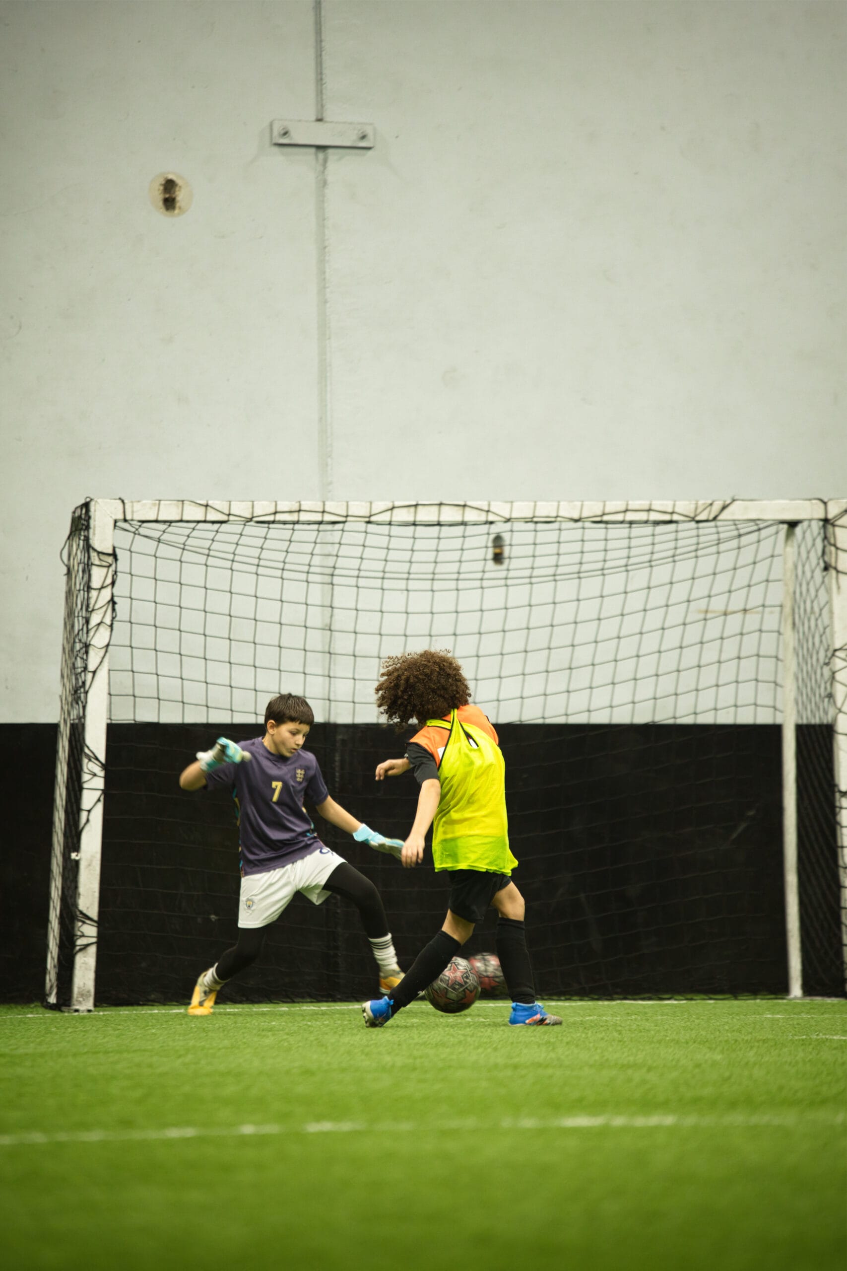 4Q8A0293 Youth soccer attacker in yellow pinnie attempting shot against goalkeeper inside indoor goal area.