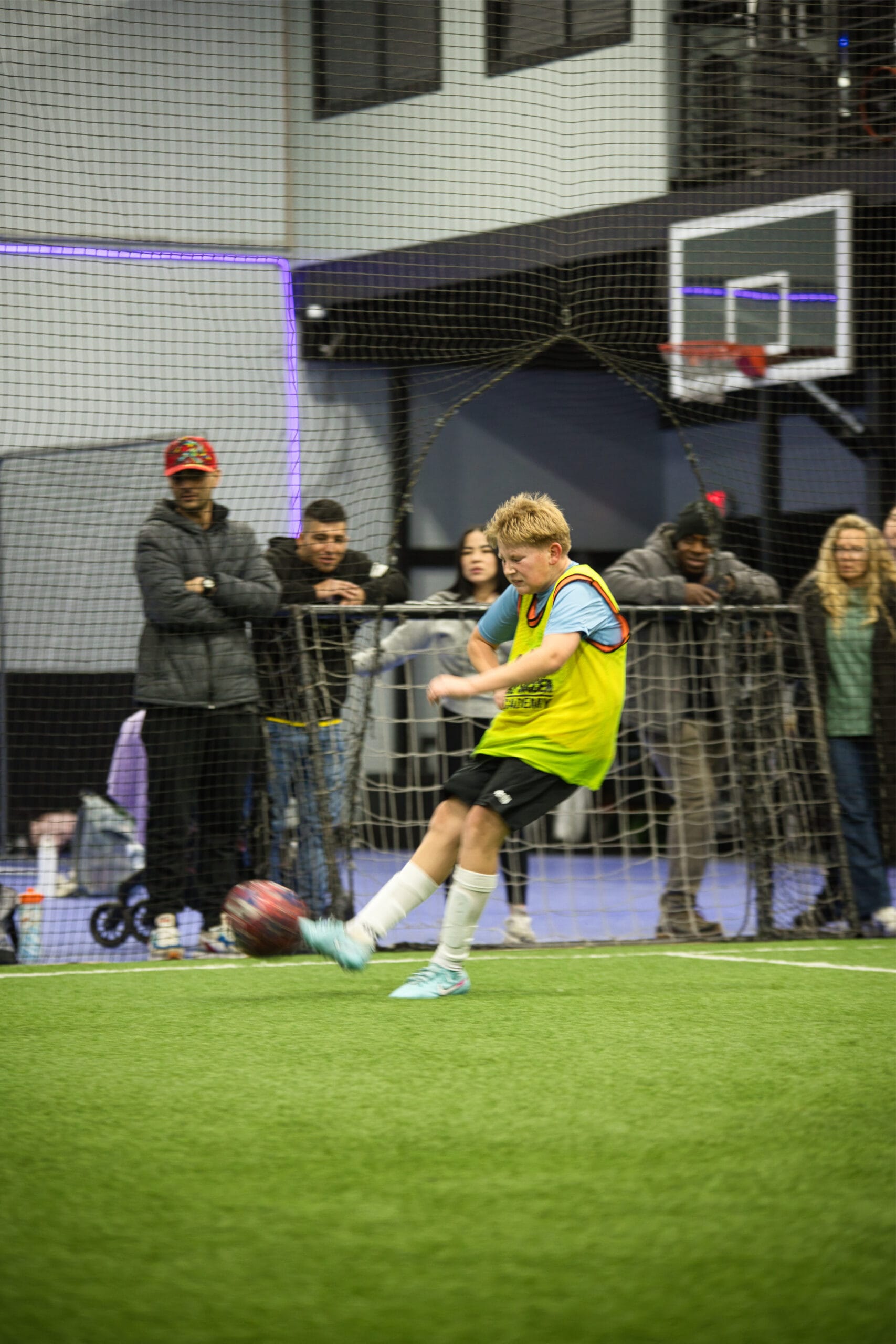 4Q8A0331 Young player striking the ball with power during indoor Pro Soccer Academy game as spectators watch behind netting.