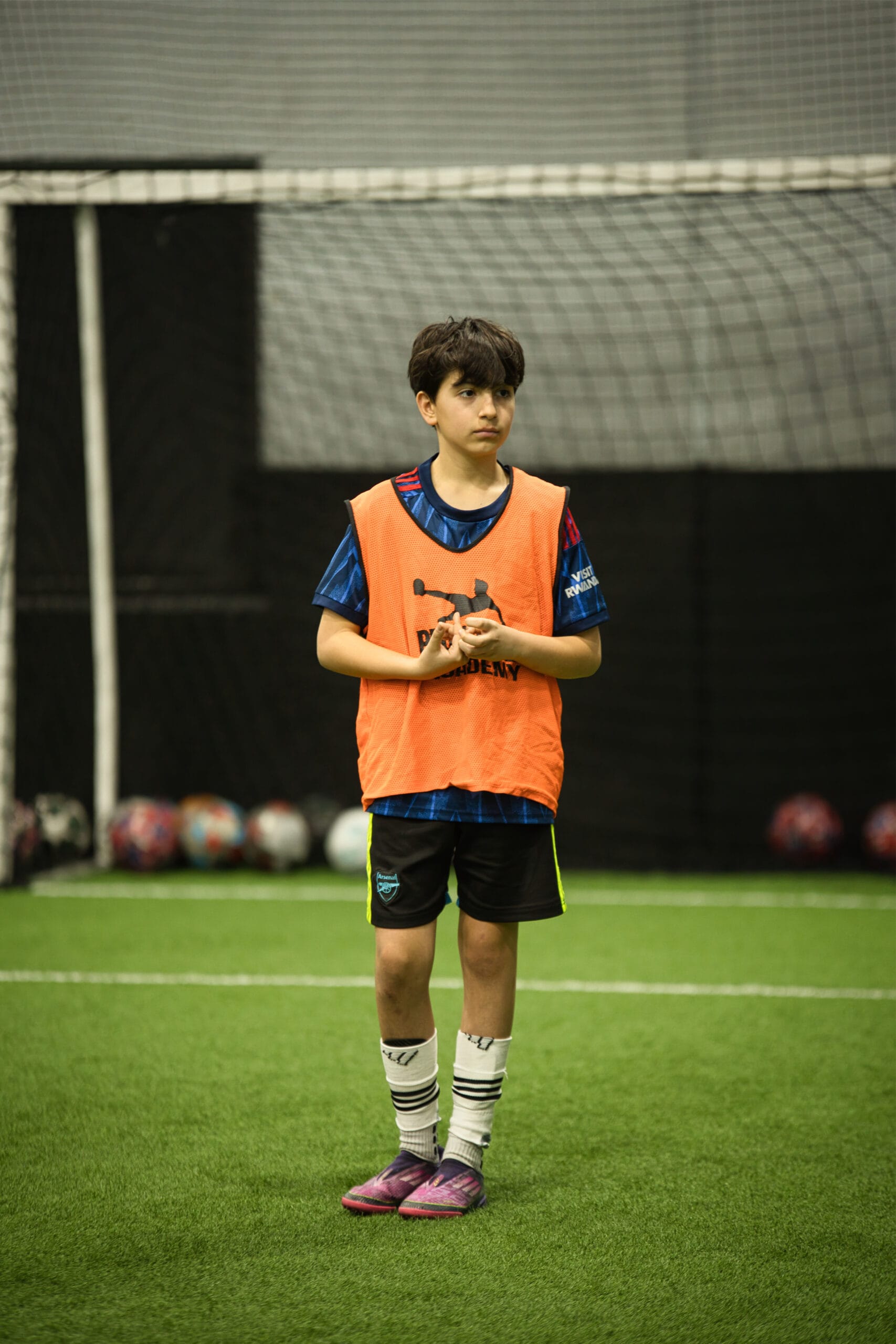 4Q8A0339 Young soccer player in orange pinnie standing focused on indoor turf in front of goal net.