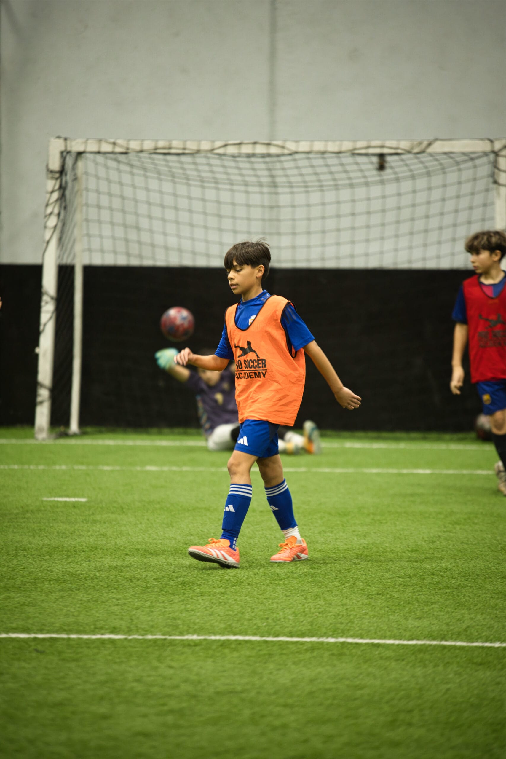 4Q8A0362 Youth player in orange pinnie jogging across field while goalkeeper dives in background during indoor scrimmage.